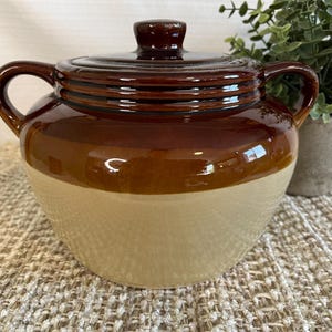 May include: A two-tone ceramic crock pot with a lid. The top and lid are a glossy brown, while the bottom half is a cream colour. It has two handles and a knob on the lid. The crock pot is sitting on a textured surface.