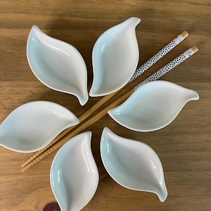 Peut inclure: Cinq coupelles en céramique blanche en forme de feuilles disposées en cercle. Deux baguettes en bois avec des motifs à pois noirs et blancs aux extrémités sont placées en diagonale au centre de la disposition des coupelles.