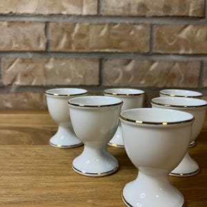 May include: A set of six white porcelain egg cups, each featuring a smooth, rounded bowl shape supported by a pedestal base. Every cup is finished with a thin, metallic gold rim along the top edge, providing a consistent and uniform appearance.