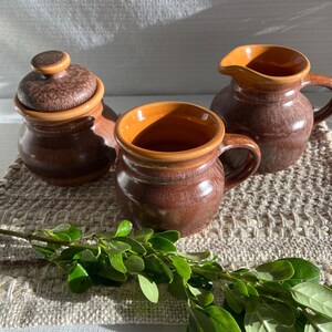 Peut inclure: Un sucrier en céramique marron avec un couvercle, une petite tasse et une crémière avec une anse. Les intérieurs sont de couleur orange contrastante. L'ensemble est sur une surface beige texturée, avec une branche de feuilles vertes.