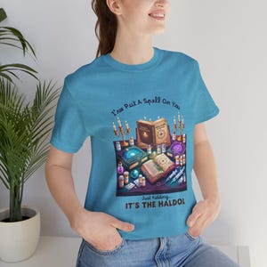 May include: A light blue t-shirt with a graphic featuring open spell books, candles, and potion bottles. The text on the shirt reads "I've Put A Spell On You" and "Just Kidding... IT'S THE HALDO!"