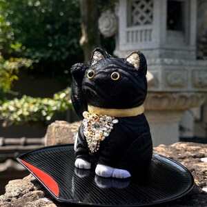 May include: Black Maneki Neko cat figurine with a raised paw, adorned with gold trim and pearl accents. The cat sits on a black tray with a red accent. The background features a stone lantern and greenery.