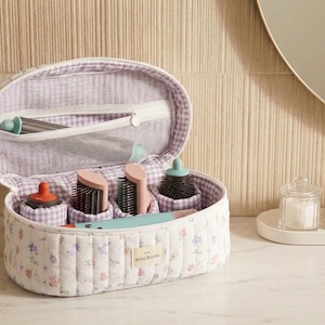 May include: An open, quilted cosmetic bag with a floral pattern, containing various brushes and tools. The bag is white with a purple and white gingham lining. A mirror and accessories are in the background.