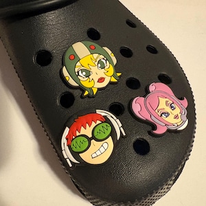 May include: A black clog shoe featuring three colorful anime-style character charms. The charms depict faces with distinct features, including one with green goggles, one with a green helmet, and one with pink hair, all attached to the shoe's holes.