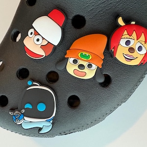 May include: Black Crocs shoe with four colorful cartoon character charms. The charms include a monkey with a red and white hat, a dog with an orange hat, a girl with red hair, and a robot with blue accents.