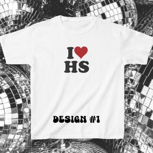 Puede incluir: Una camiseta blanca de manga corta con un estampado gr&aacute;fico en el pecho. El dise&ntilde;o incluye un s&iacute;mbolo de coraz&oacute;n rojo entre la letra I y las letras HS en negro. Debajo, el texto DESIGN #1 est&aacute; impreso en una fuente negra en negrita.