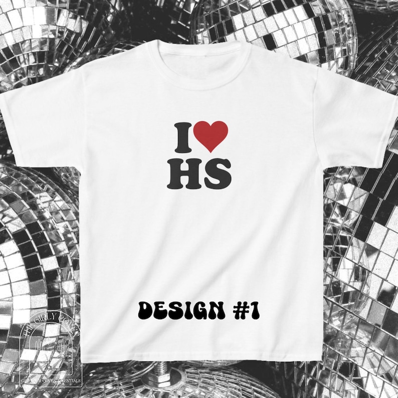 Puede incluir: Una camiseta blanca de manga corta con un estampado gr&aacute;fico en el pecho. El dise&ntilde;o incluye un s&iacute;mbolo de coraz&oacute;n rojo entre la letra I y las letras HS en negro. Debajo, el texto DESIGN #1 est&aacute; impreso en una fuente negra en negrita.