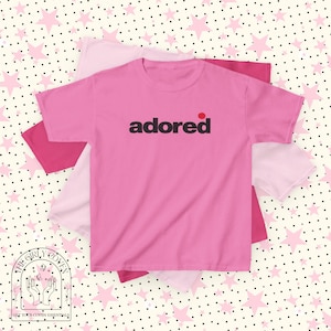 May include: A bright pink short-sleeved t-shirt featuring the word "adored" printed in black lowercase letters across the chest, with a small red dot positioned above the letter "d". The shirt is layered over other pink t-shirts.