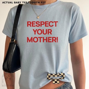 Puede incluir: Una camiseta de manga corta de color azul claro con el texto en rojo RESPECT YOUR MOTHER! impreso en la parte delantera. La camiseta se combina con un bolso de hombro negro de acabado texturizado y una pinza de pelo a cuadros blancos y negros.