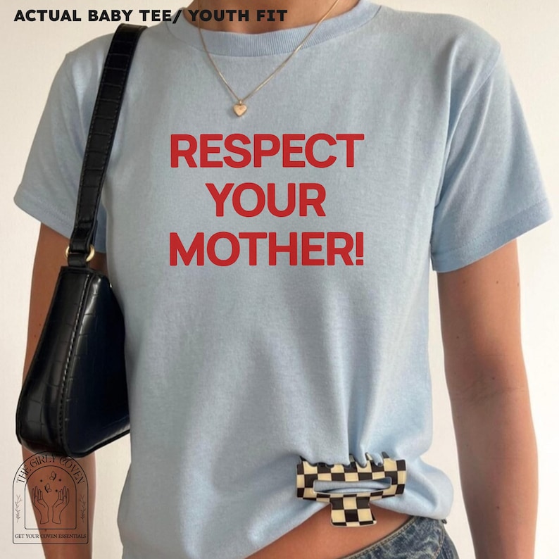Puede incluir: Una camiseta de manga corta de color azul claro con el texto en rojo RESPECT YOUR MOTHER! impreso en la parte delantera. La camiseta se combina con un bolso de hombro negro de acabado texturizado y una pinza de pelo a cuadros blancos y negros.