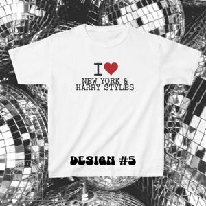 Puede incluir: Una camiseta blanca de manga corta con el texto negro I, un s&iacute;mbolo de coraz&oacute;n rojo y las palabras NEW YORK & HARRY STYLES. Debajo del gr&aacute;fico, el texto DESIGN #5 est&aacute; impreso en una fuente negra, negrita y redondeada en la parte frontal.