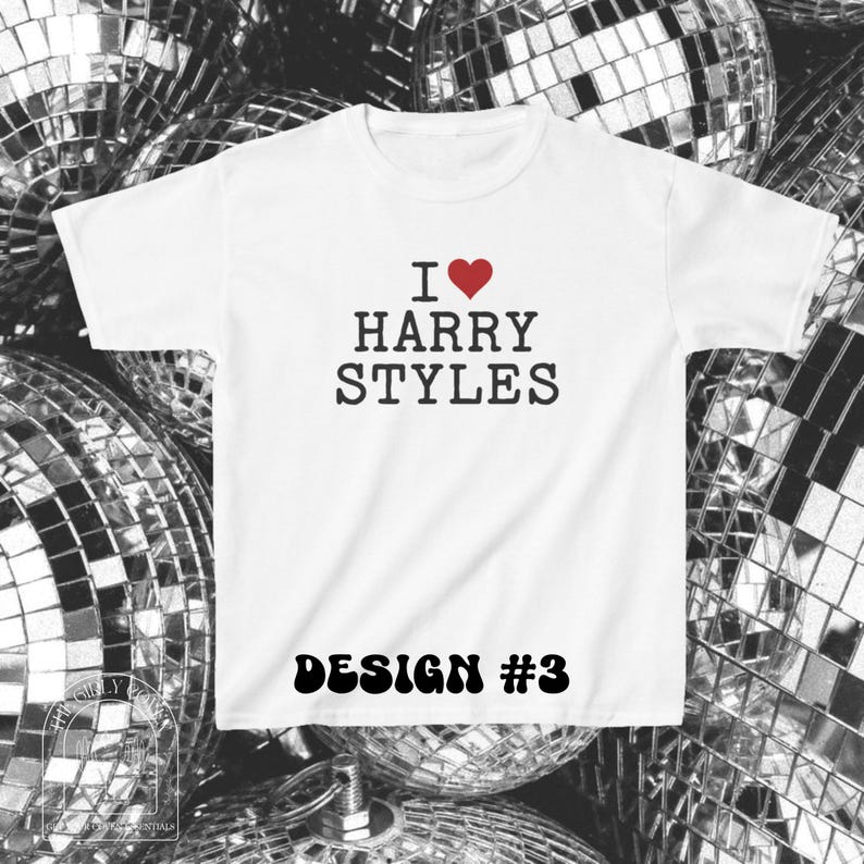 Puede incluir: Una camiseta blanca de manga corta con el texto negro I, un s&iacute;mbolo de coraz&oacute;n rojo y las palabras HARRY STYLES. Debajo del texto, las palabras DESIGN #3 est&aacute;n impresas en una fuente negra, negrita y redondeada. La camiseta tiene cuello redondo.