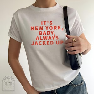 Puede incluir: Una camiseta blanca de manga corta con el texto rojo IT'S NEW YORK, BABY, ALWAYS JACKED UP impreso en la parte delantera. La camiseta se combina con un bolso de hombro negro con acabado texturizado y anillos dorados en los dedos.