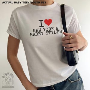 Puede incluir: Una camiseta blanca de manga corta con cuello redondo. La parte delantera presenta un gr&aacute;fico con un coraz&oacute;n rojo y el texto negro I LOVE NEW YORK & HARRY STYLES. La camiseta se lleva con vaqueros azules y un bolso de hombro negro.