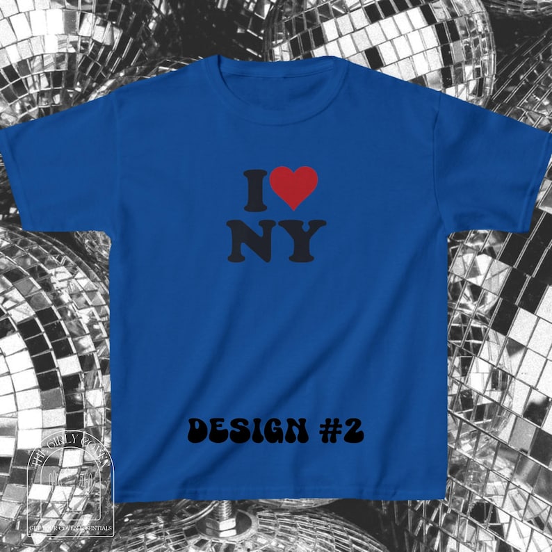 Puede incluir: Una camiseta de manga corta color azul real con el texto negro I, un s&iacute;mbolo de coraz&oacute;n rojo y el texto negro NY. Debajo del gr&aacute;fico, el texto DESIGN #2 est&aacute; impreso en negro. La camiseta tiene cuello redondo y un corte est&aacute;ndar.