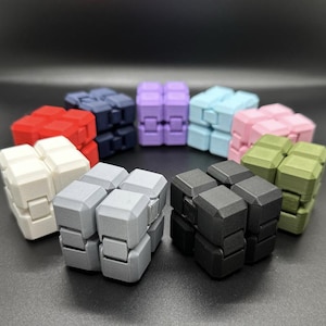May include: A collection of colorful fidget cubes in various shades, including red, white, gray, black, navy, purple, light blue, pink, and olive green. The cubes are arranged in a circular pattern, showcasing their geometric design and multiple color options.