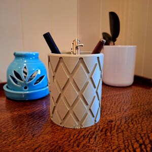 May include: A cylindrical light gray pen holder featuring a repeating diamond-shaped geometric pattern on its exterior surface. The container holds three writing instruments, including a black pen, a silver metal pen, and a dark brown wooden pen.