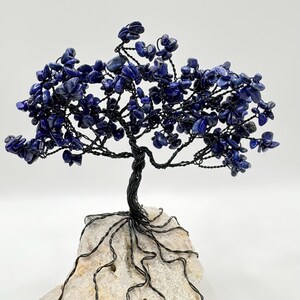May include: A handcrafted wire tree sculpture with a dark, twisted trunk and branches. The tree's foliage is composed of small, deep blue gemstones. The tree is mounted on a light-colored, textured stone base.