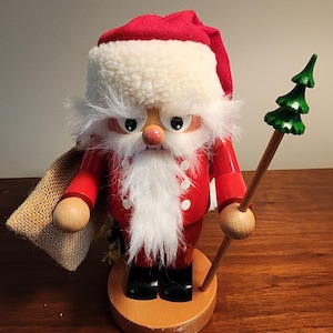 May include: A wooden Santa Claus figurine with a red coat and hat, white beard and fur trim, and black boots. He holds a green tree on a wooden staff and a burlap sack. The figurine is on a round wooden base.