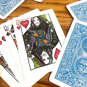 May include: A custom playing card featuring a Queen of Hearts with a portrait of a person labelled Kate, wearing a green and orange patterned garment. The card includes a red heart symbol and the letter Q in the corner, with other blue-backed cards.