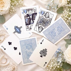 May include: A set of custom playing cards featuring a king and queen with illustrated faces, an ace of diamonds with a blue ring graphic, an ace of spades with a cake graphic, a three of clubs, and blue card backs with floral text designs.