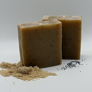May include: Two rectangular bars of handmade soap, light brown with dark specks. A small pile of brown granules and scattered black seeds are next to the soap. The soap has a natural, rustic appearance.