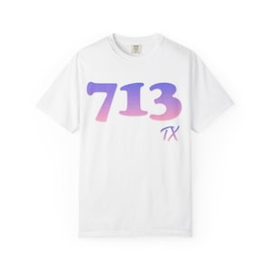May include: White t-shirt with a crew neck featuring the number "713" in a gradient of purple and pink. The letters "TX" are below the number in a matching gradient. The shirt is made of a soft material.