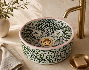 May include: A round ceramic vessel sink featuring a white base with intricate green floral and geometric patterns, a soft pink rim, and a central gold-toned drain, paired with a tall, brushed gold-toned metal faucet.