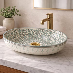 Puede incluir: Lavabo ovalado de cerámica con un patrón decorativo floral y de volutas en verde azulado y blanco. El lavabo tiene un desagüe dorado y está colocado sobre una encimera de mármol. Un grifo dorado está montado en la pared detrás del lavabo.