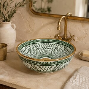 May include: A decorative ceramic sink with a green and white geometric pattern and gold trim. The sink is round and sits on a light-colored countertop. A gold faucet and mirror are in the background, suggesting a bathroom setting.