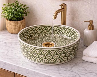 May include: A decorative, round bathroom sink with a green and white scalloped pattern. The sink is set on a marble countertop with a gold faucet. A small potted plant and a soap dispenser are also visible.