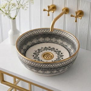 May include: A decorative ceramic sink with a black and white geometric pattern and a gold rim. The sink is round and sits on a white surface. A gold faucet and fixtures are mounted on the wall behind the sink.