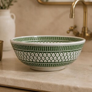 May include: A white ceramic bowl with a green geometric pattern. The bowl features a repeating design of small, connected teardrop shapes and a border of green squares. The bowl is sitting on a light-colored countertop.