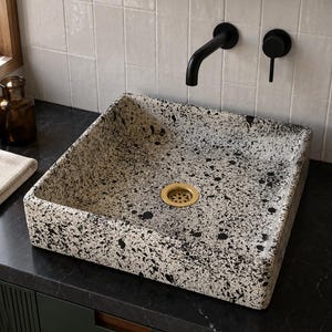 May include: A square vessel sink featuring a speckled pattern of black and white paint splatters. The sink includes a circular brass-toned drain cover centered at the bottom, contrasting with the monochromatic speckled finish of the basin.