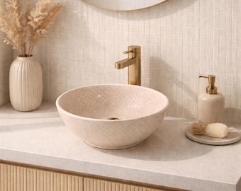May include: A bathroom vanity with a light beige ceramic vessel sink featuring a crackle pattern. A gold faucet, mirror, and soap dispenser complement the neutral color scheme. A vase with dried plants and a wooden cabinet complete the design.
