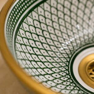 May include: Close-up of a decorative ceramic sink basin with a white and green geometric pattern. The basin has a gold rim and a gold drain. The design features a repeating pattern of diamond shapes with small dots.