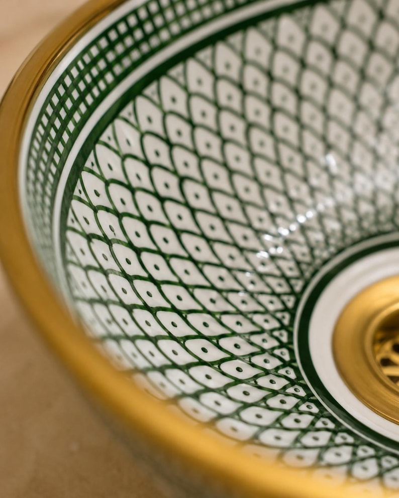 May include: Close-up of a decorative ceramic sink basin with a white and green geometric pattern. The basin has a gold rim and a gold drain. The design features a repeating pattern of diamond shapes with small dots.