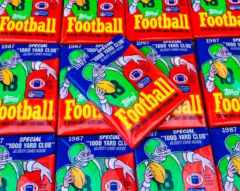 May include: A close-up of a stack of 1987 Topps Football trading cards featuring a football player wearing a green and white uniform. The cards are red, white, and blue with the text "1000 Yard Club" and "Special" printed on them.