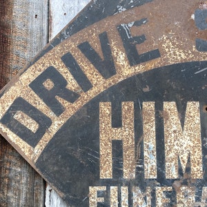 May include: Aged metal sign featuring the text "DRIVE SAFE" and "HIMM" in white lettering against a black backdrop. The sign displays a distressed, rusty finish, indicating its age. It is mounted on a weathered wooden surface, adding to its vintage appeal.