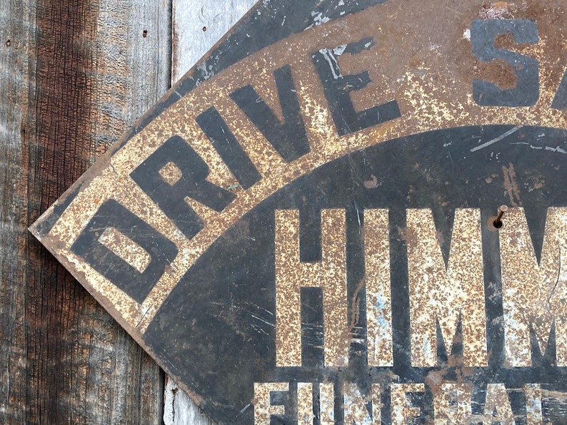 May include: Aged metal sign featuring the text "DRIVE SAFE" and "HIMM" in white lettering against a black backdrop. The sign displays a distressed, rusty finish, indicating its age. It is mounted on a weathered wooden surface, adding to its vintage appeal.