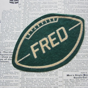 May include: A green and cream-colored patch in the shape of a football, with the word "FRED" in cream-colored block letters. The patch is on a vintage paper background.