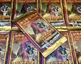 Puede incluir: Primer plano de una pila de paquetes de cartas de fútbol americano Dynagon Prism 1997 sin abrir. Los paquetes son dorados y rojos con las palabras "Dynagon Prism" y "1997 NFL Football Cards" impresas en ellos.