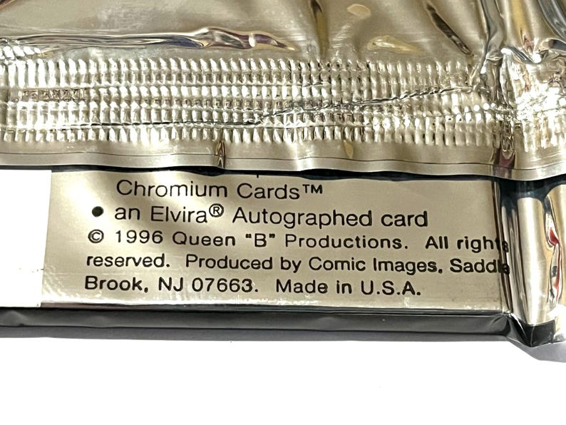Puede incluir: Primer plano de un paquete sellado de "Chromium Cards" con una tarjeta autografiada de Elvira. El embalaje plateado y met&aacute;lico tiene texto que indica la producci&oacute;n de la tarjeta por Comic Images en 1996, fabricado en EE. UU.