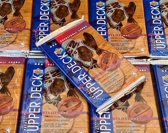 Può includere: Più pacchetti di carte di basket NBA Upper Deck. I pacchetti sono blu, rossi e oro, con immagini di giocatori di basket e palloni. Il testo "Upper Deck" e "Series One" sono visibili.
