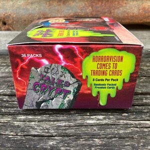 Puede incluir: Una caja de cartas coleccionables de Tales from the Crypt. La caja es roja y verde con una imagen de una l&aacute;pida que dice "Tales from the Crypt". La caja dice "Horrorvision Comes to Trading Cards" y "36 Packs".