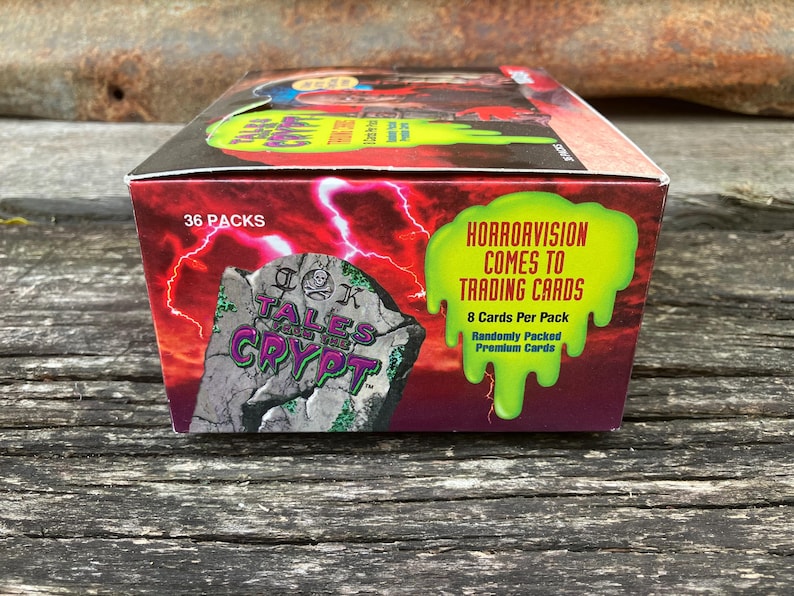 Puede incluir: Una caja de cartas coleccionables de Tales from the Crypt. La caja es roja y verde con una imagen de una l&aacute;pida que dice "Tales from the Crypt". La caja dice "Horrorvision Comes to Trading Cards" y "36 Packs".