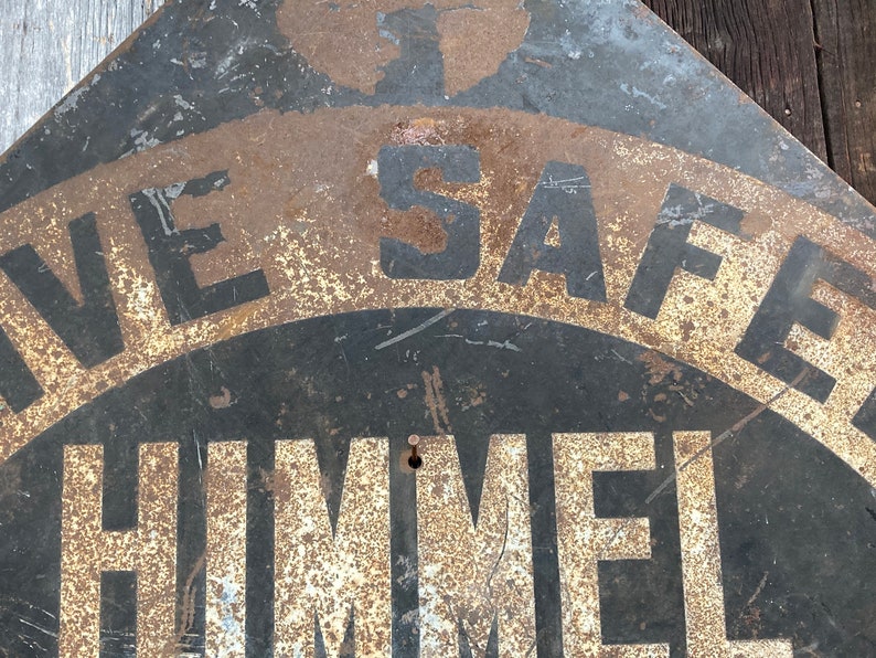 May include: A weathered metal sign featuring the text "LIVE SAFE" and "HIMMEL" in black and white. The sign has a distressed, rusty finish, indicating it's an older piece. The sign's design suggests it's an antique or vintage item.
