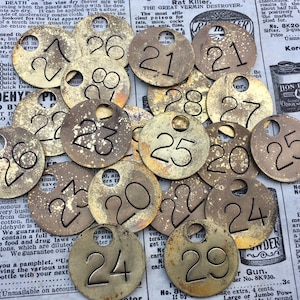 May include: A collection of 16 brass numbered tags, each with a hole in the center. The numbers range from 1 to 29. The tags are arranged in a random pattern on a newspaper background.