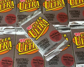 Puede incluir: Primer plano de una pila de tarjetas de fútbol de colección 1992 Fleer Ultra. Las tarjetas presentan un fondo plateado con un gráfico de fútbol amarillo y naranja y el texto "92 Fleer Ultra". Las tarjetas también presentan los nombres de los jugadores de fútbol Reggie White y Chris Miller.