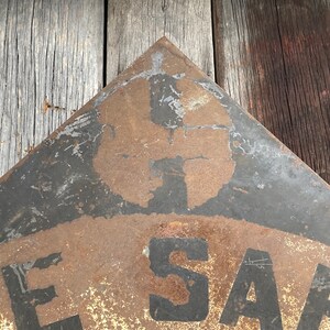 May include: A weathered, triangular metal sign with the word "SAFE" visible. The sign has a black border and a faded, rusty appearance, suggesting age and outdoor exposure. The background is a wooden surface.